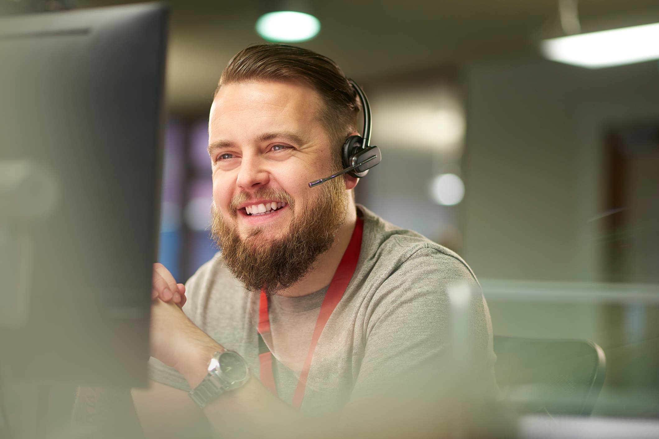 A bearded help‑desk specialist wearing a headset and lanyard smiles confidently in a modern office, exemplifying professional managed IT services support.